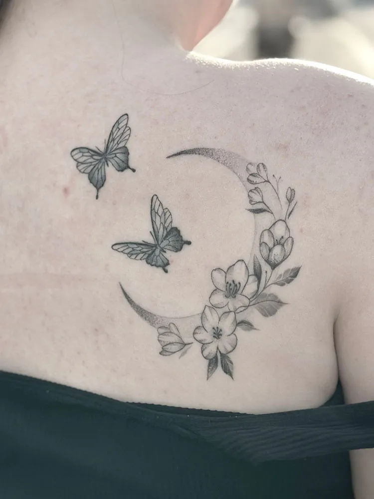 Fineline tatovering af måne med blomster og sommerfugle på ryggen