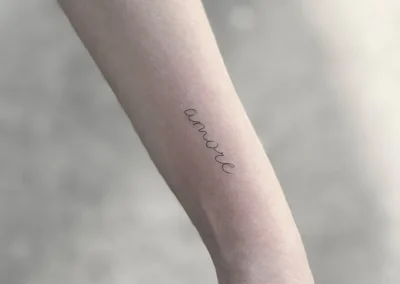 Fineline-tatovering med ordet amore i håndskrift på underarmen