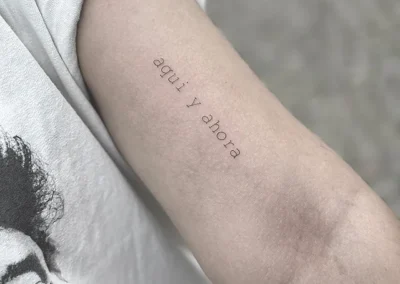 Fineline-tatovering med teksten aqui y ahora på overarmen