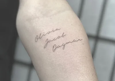 Fineline-tatovering med tre børnenavne i håndskrift på overarmen