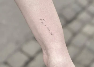 Fineline-tatovering med ordet Farmor i håndskrift på underarmen