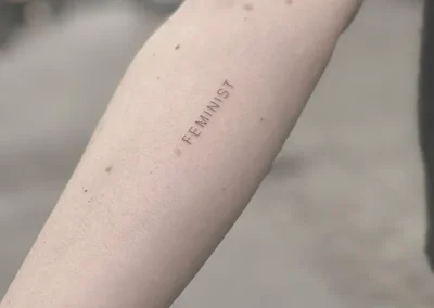 Fineline-tatovering med ordet FEMINIST i små versaler på underarmen