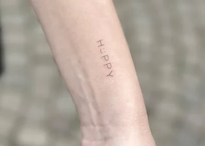 Fineline-tatovering med ordet HAPPY i små versaler på underarmen