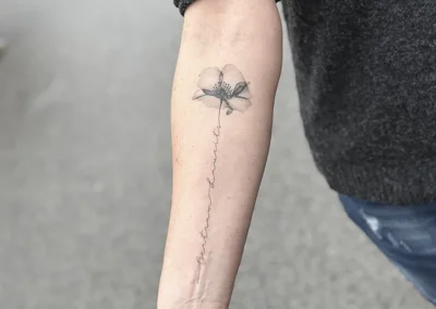 Helet fineline-tatovering af en blomst med lang stilk ned ad underarmen