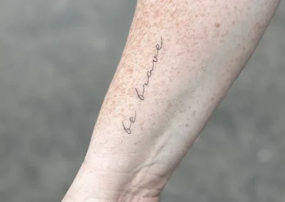 Helet fineline-tatovering med ordene be brave i håndskrift på underarmen