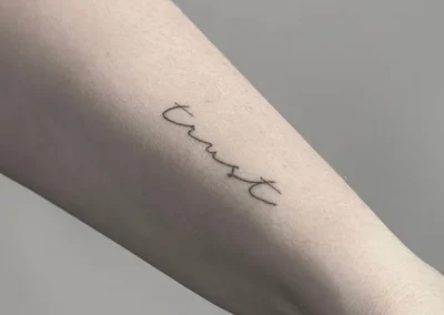 Helet fineline-tatovering med ordet trust i håndskrift på underarmen