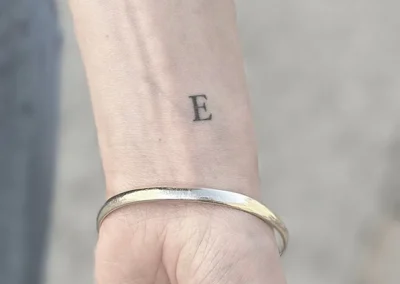 Helet fineline-tatovering med bogstavet E på håndleddet