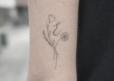 Helet fineline-tatovering med blomster og initialet E på overarmen