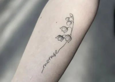 Helet fineline-tatovering med liljekonvaller og navnet Monse på underarmen