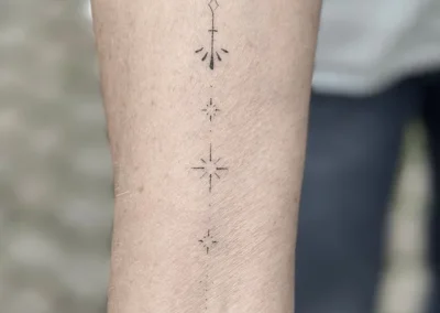 Helet fineline-tatovering med et ornament og stjerner i en lodret linje på underarmen