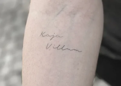 Helet fineline-tatovering med navnene Kaja og Villum i håndskrift på overarmen