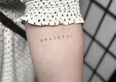 Helet fineline-tatovering med ordet GRATEFUL i små versaler på overarmen