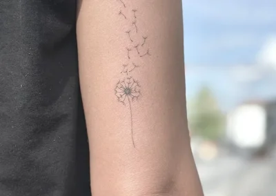 Fineline-tatovering af en mælkebøtte med frø der flyver væk, på overarmen