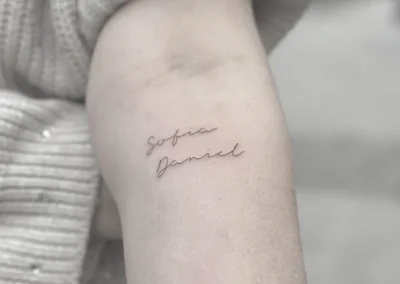 Fineline-tatovering med to navne i håndskrift på overarmen